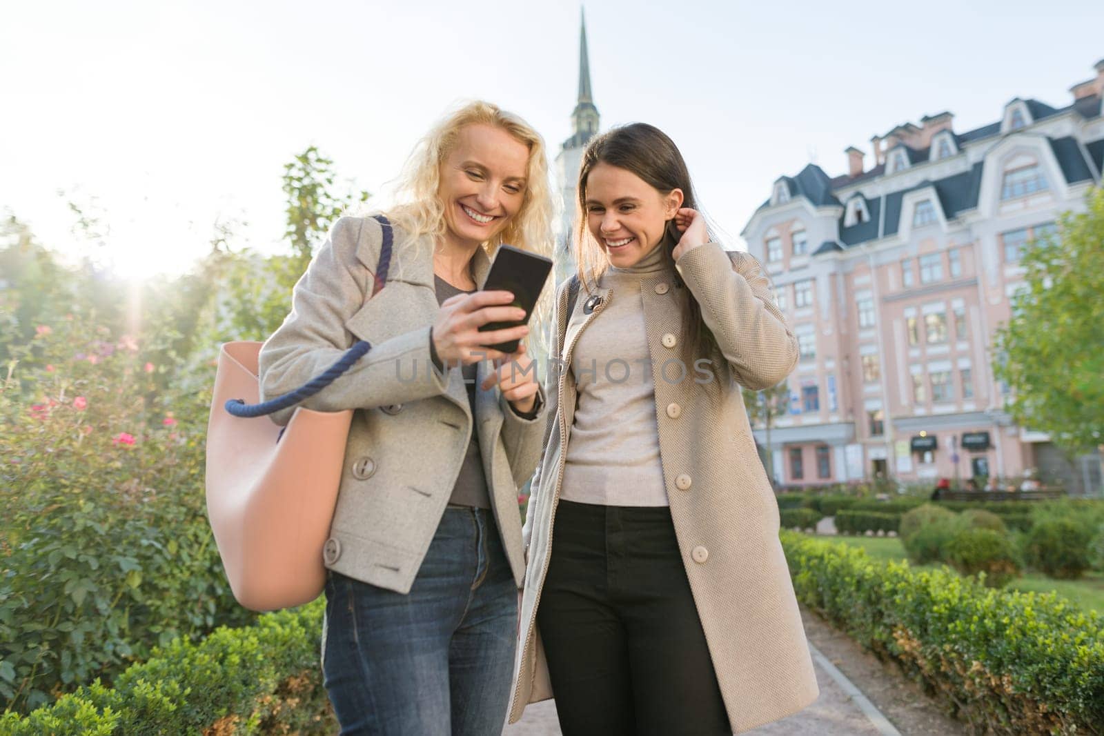Two young women having fun, looking at the smartphone laughing, sunny autumn day, city background, golden hour.