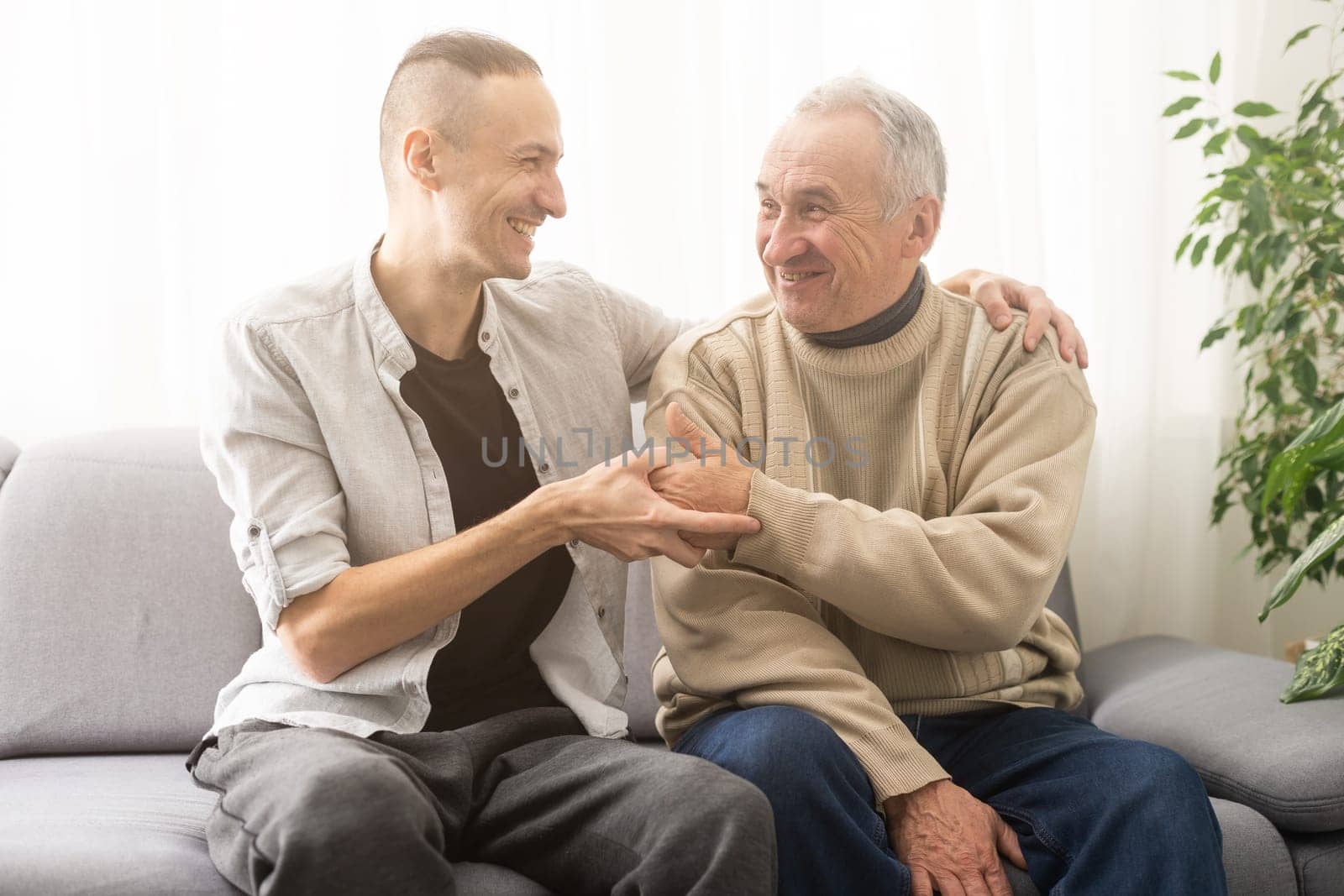 Happy two generations male family old senior mature father and smiling young adult grown son enjoying talking chatting bonding relaxing having friendly positive conversation sit on sofa at home. by Andelov13
