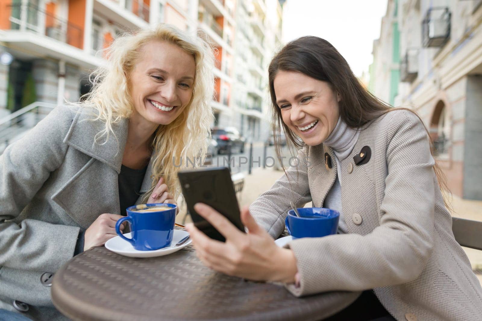Two young smiling women having fun in outdoor cafe. Urban autumn background.