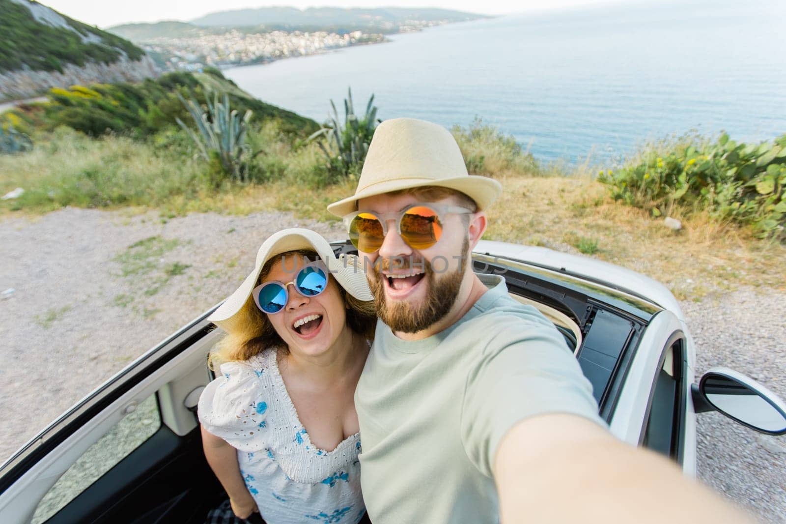 Happy beautiful couple in love taking a selfie portrait driving a convertible car on the road at vacation. Rental cars and vacation by Satura86