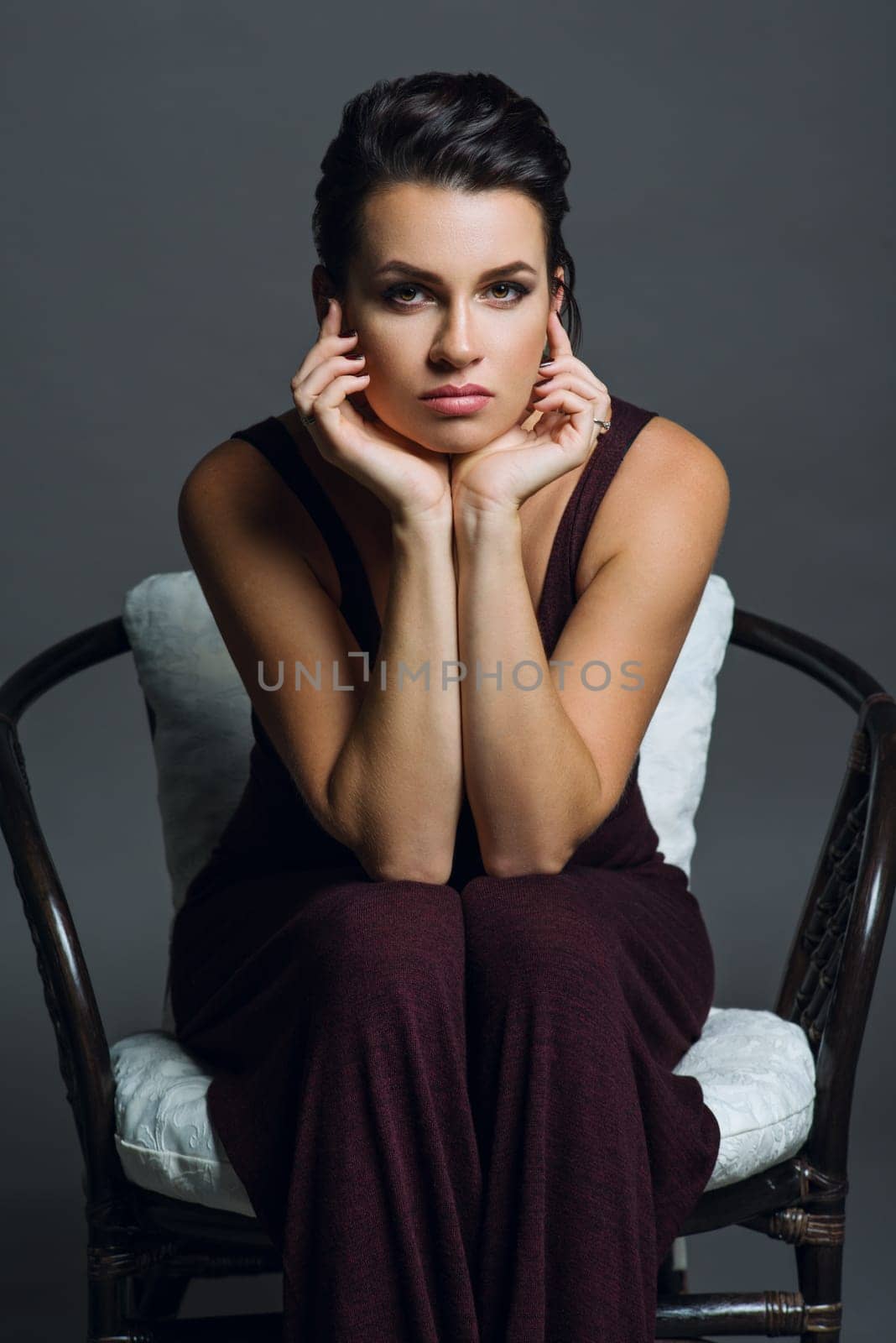 Fashionable beautiful young woman with hairstyle and makeup posing on a dark background. Female in trendy knitted overalls on a wicker chair