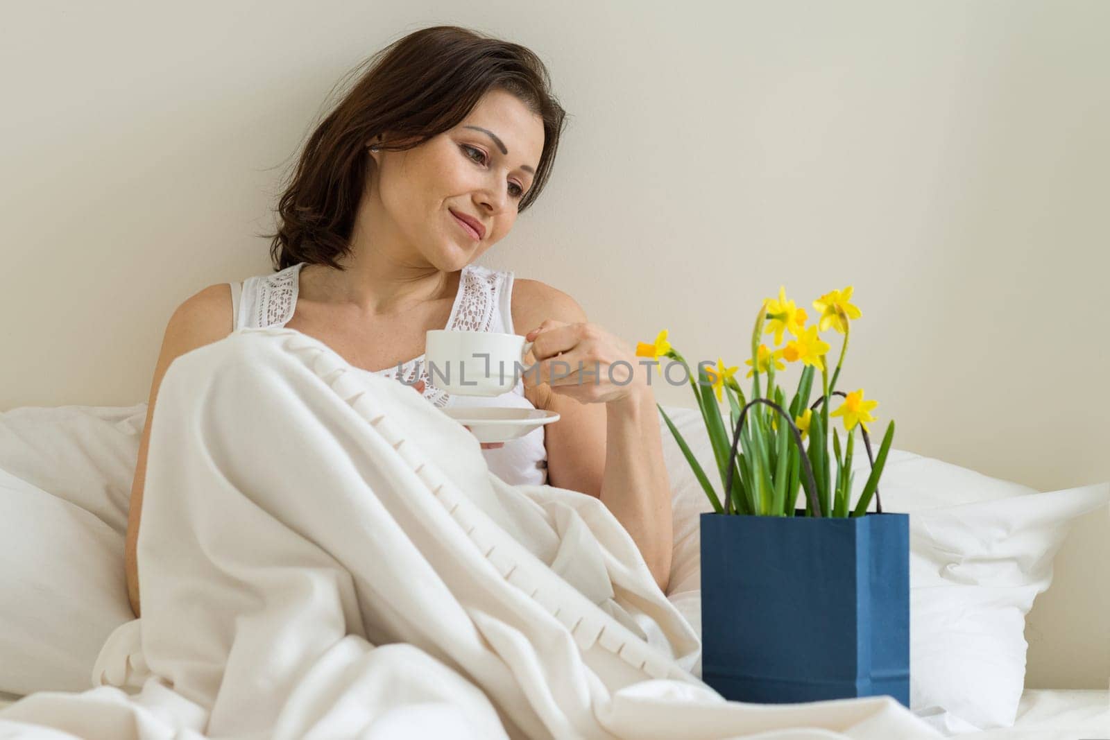 Middle aged woman is pleased with gift, bouquet of flowers sitting in the morning in bed with a cup of coffee. Emotion of happiness, joy, surprise