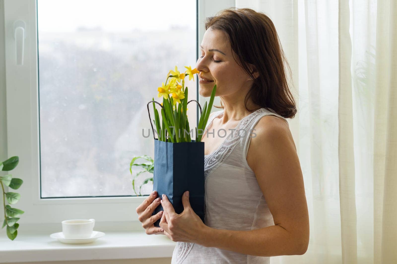 Mature woman holding bouquet of yellow spring flowers, enjoys flowers. Picture of the house, in the morning near the window