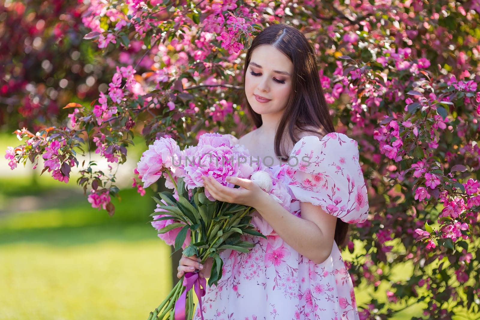 A pretty brunette in a light pink dress, with a bouquet of pink peonies, stands near pink blooming apple trees,in the garden . Copy space.
