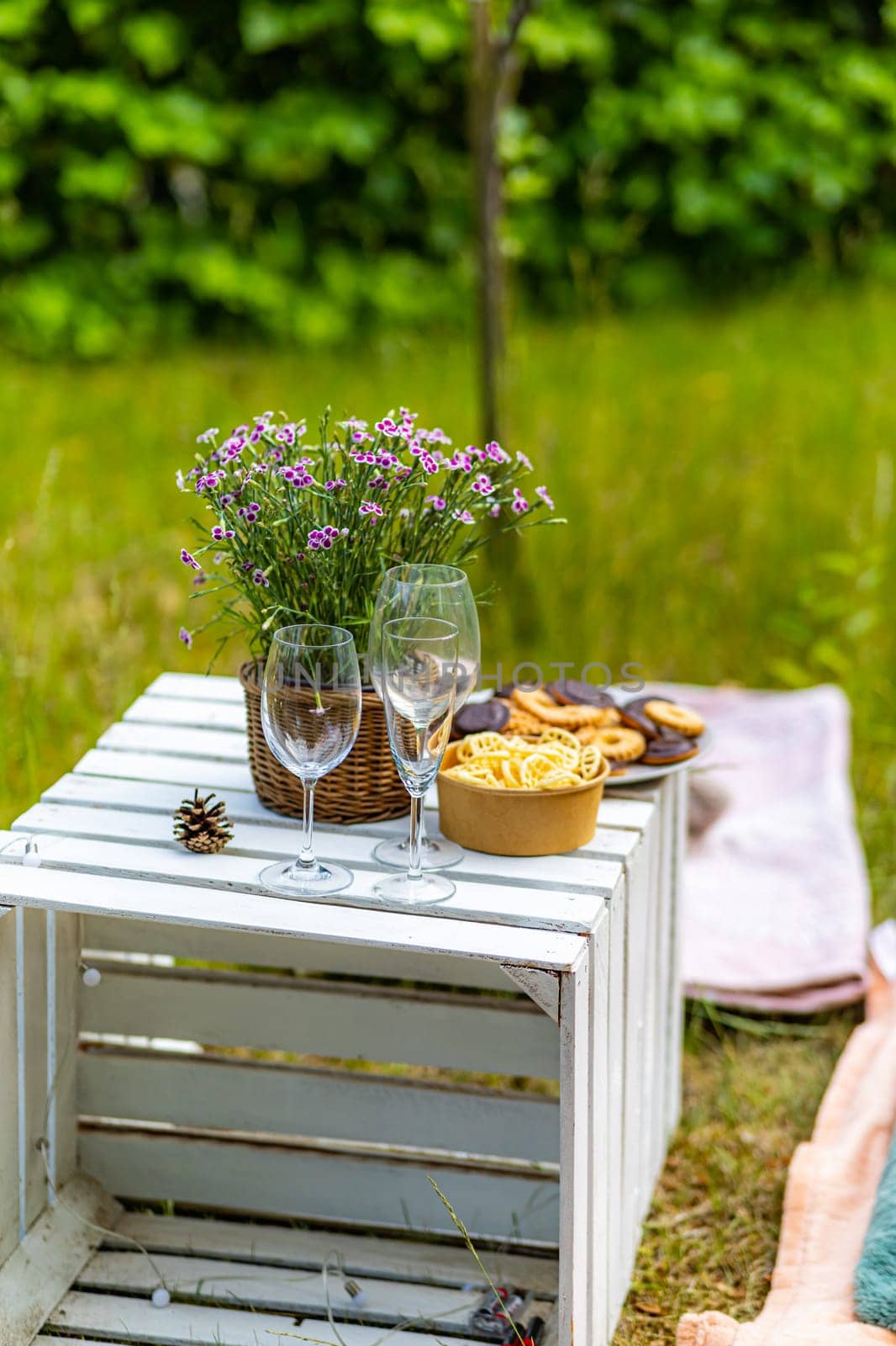 A green garden with a white wooden crate on which there are glasses, flowers and snacks in bowls by Wierzchu