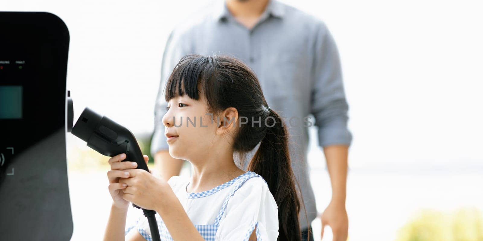 Focus progressive young girl holding EV charger device from home electric charging station for renewable energy powered car with blur asian man background. Sustainable green energy technology concept.