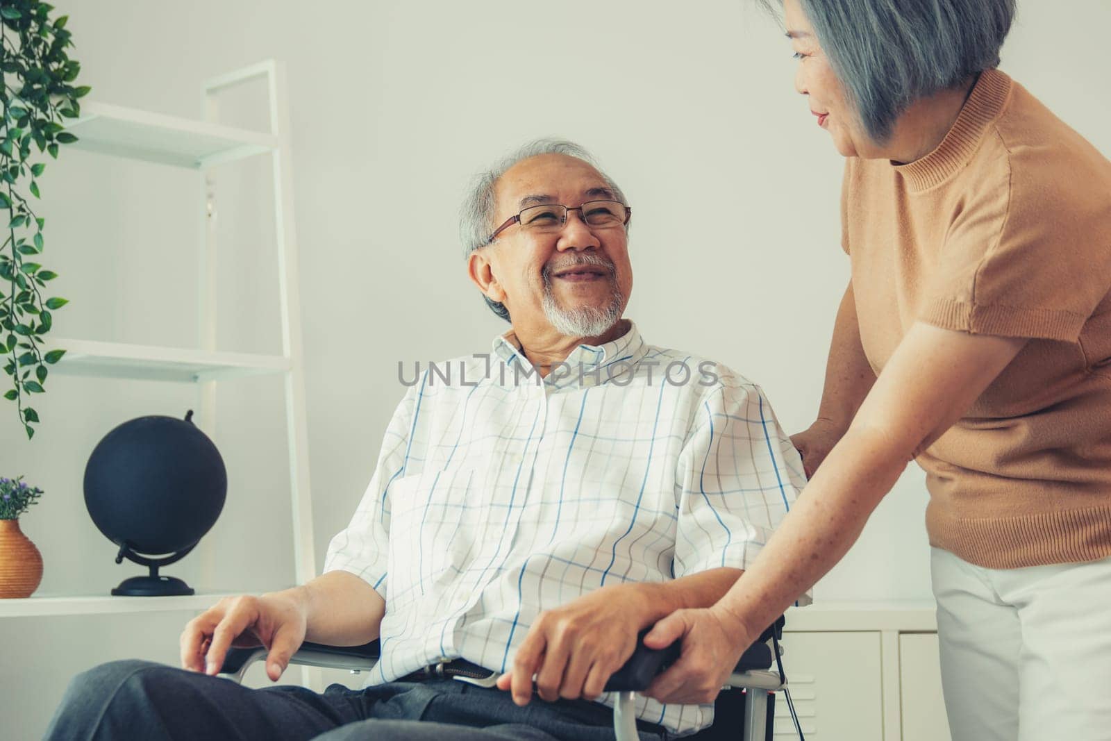 Senior wife giving support to her husband in his wheelchair with love, contented pensioner life. A senior couple is understanding and smiling at each other.