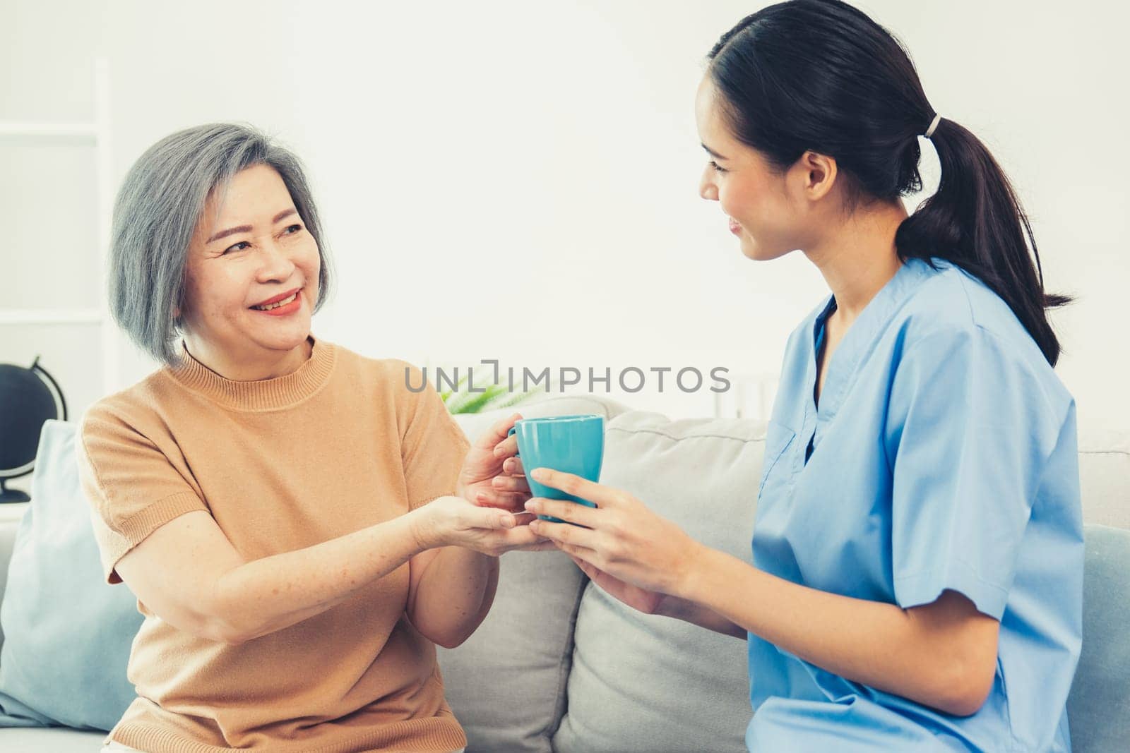 Female care taker serving her contented senior patient with a cup of coffee at home, smiling to each other. Medical care for pensioners, Home health care service.