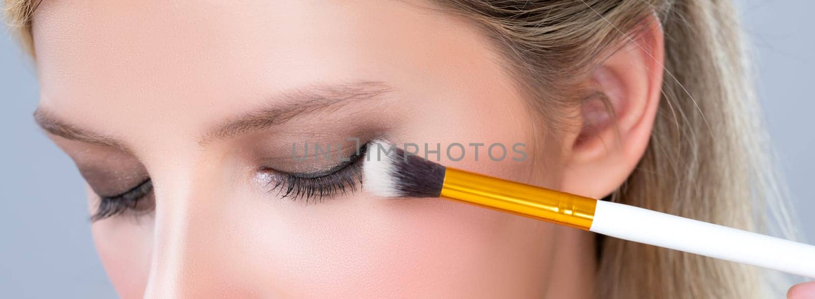 Closeup beautiful girl with flawless applying alluring eye shadow makeup with eyeliner brush. Cosmetic facial painting process on lovely young woman with perfect clean skin on isolated background.