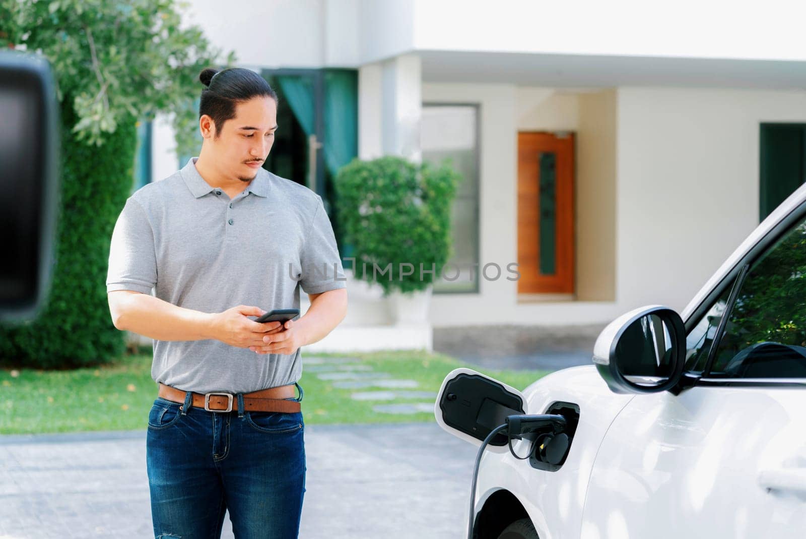 Progressive asian man install cable plug to his electric car with home charging station in the backyard. Concept use of electric vehicles in a progressive lifestyle contributes to clean environment.