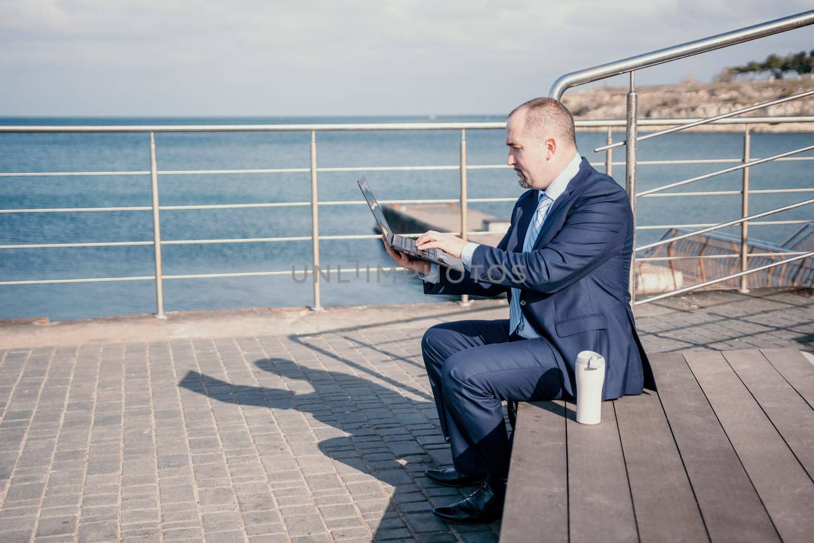 Confident middle age businessman working remotely online, typing on a laptop keyboard while sitting on a beach at sunset. Working remotely on vacation, running an online business from a distance. by panophotograph