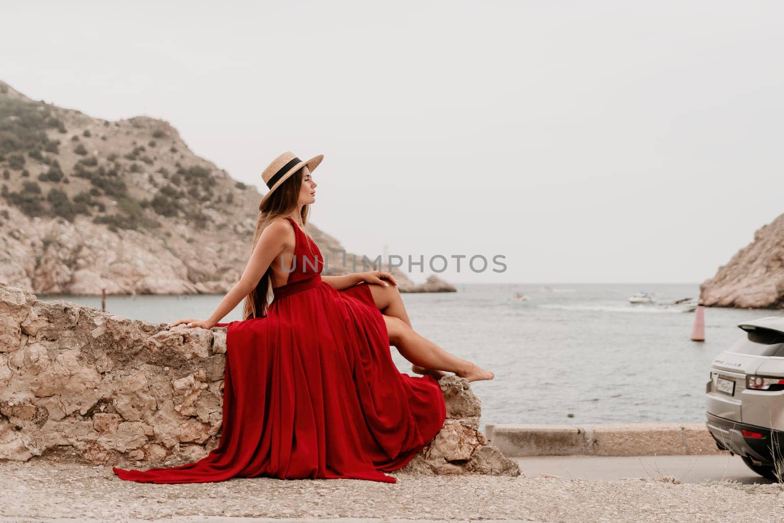 Side view a Young beautiful sensual woman in a red long dress posing on a volcanic rock high above the sea during sunset. Girl on the nature on overcast sky background. Fashion photo