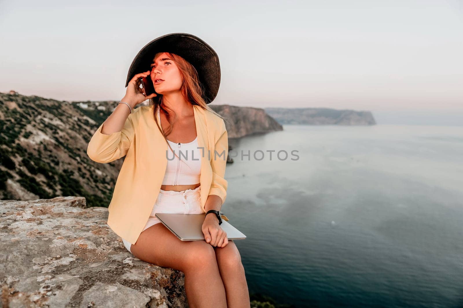 Successful business woman in yellow hat working on laptop by the sea. Pretty lady typing on computer at summer day outdoors. Freelance, travel and holidays concept.