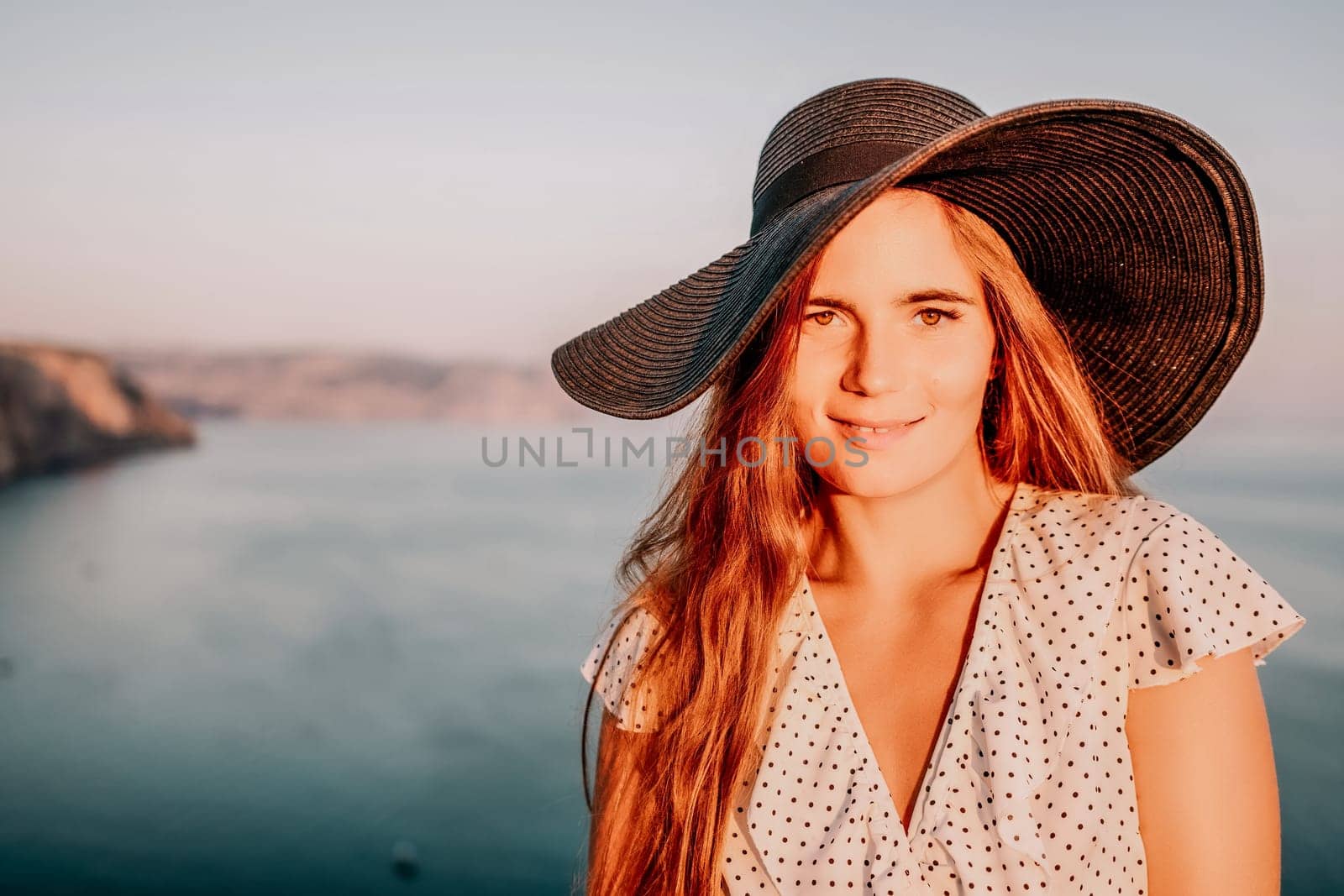 Portrait of happy young woman wearing summer black hat with large brim at beach on sunset. Closeup face of attractive girl with black straw hat. Happy young woman smiling and looking at camera at sea