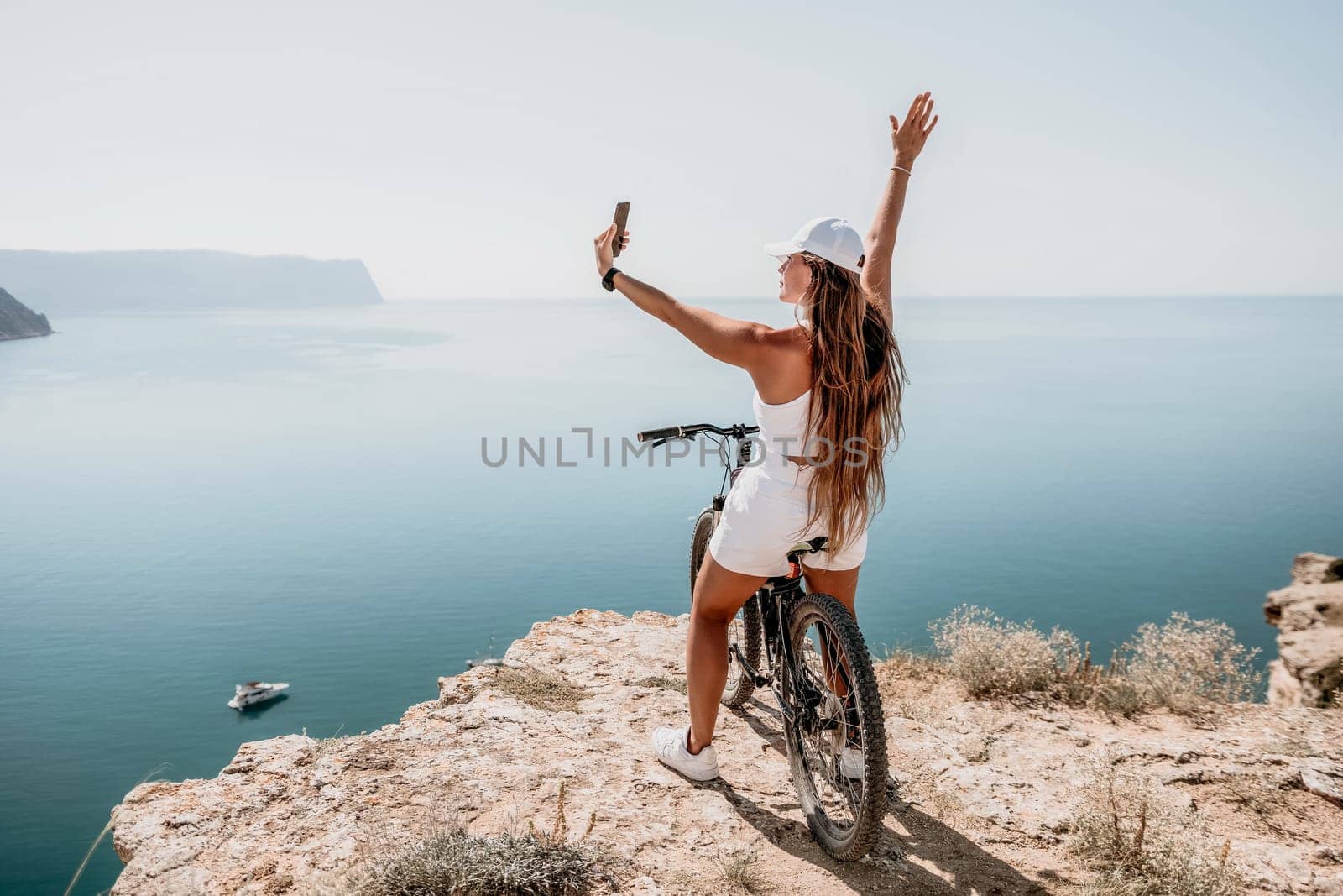 A woman cyclist on a mountain bike looking at the landscape of mountains and sea. Rear view of cyclist woman standing in front to the sea with outstretched arms. Freedom and healthy lifestyle concept