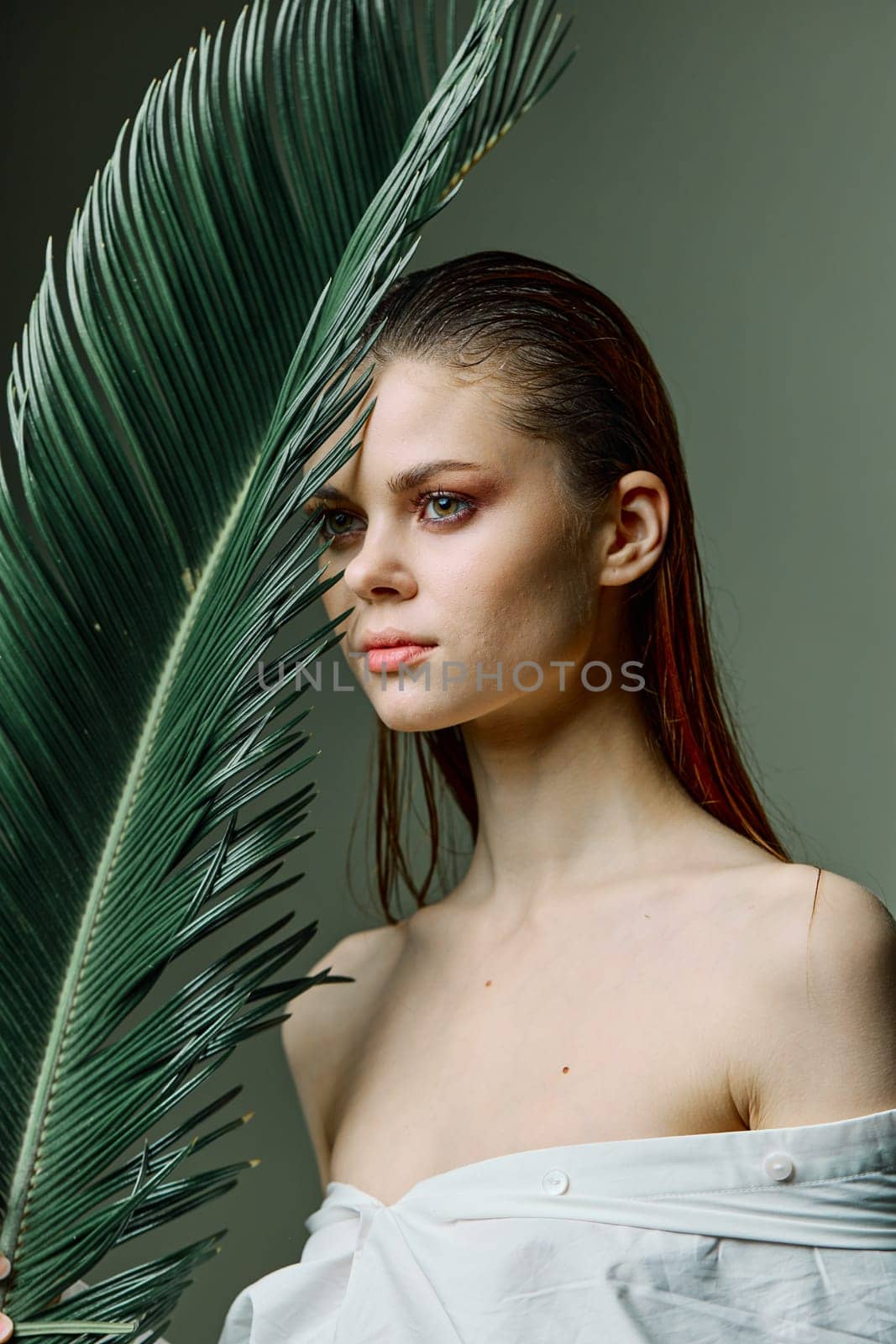 vertical photo portrait of an elegant woman with her hair slicked back, standing with a palm leaf holding it near her face. High quality photo