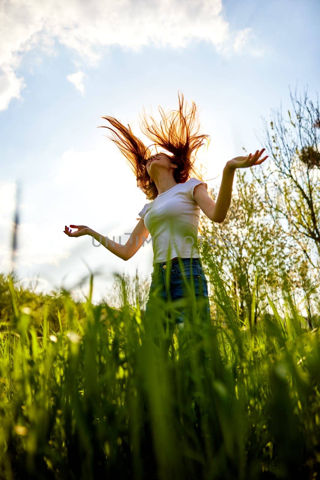a happy woman in summer clothes stands in a field lit from behind and with her hair flying in the air by Vichizh