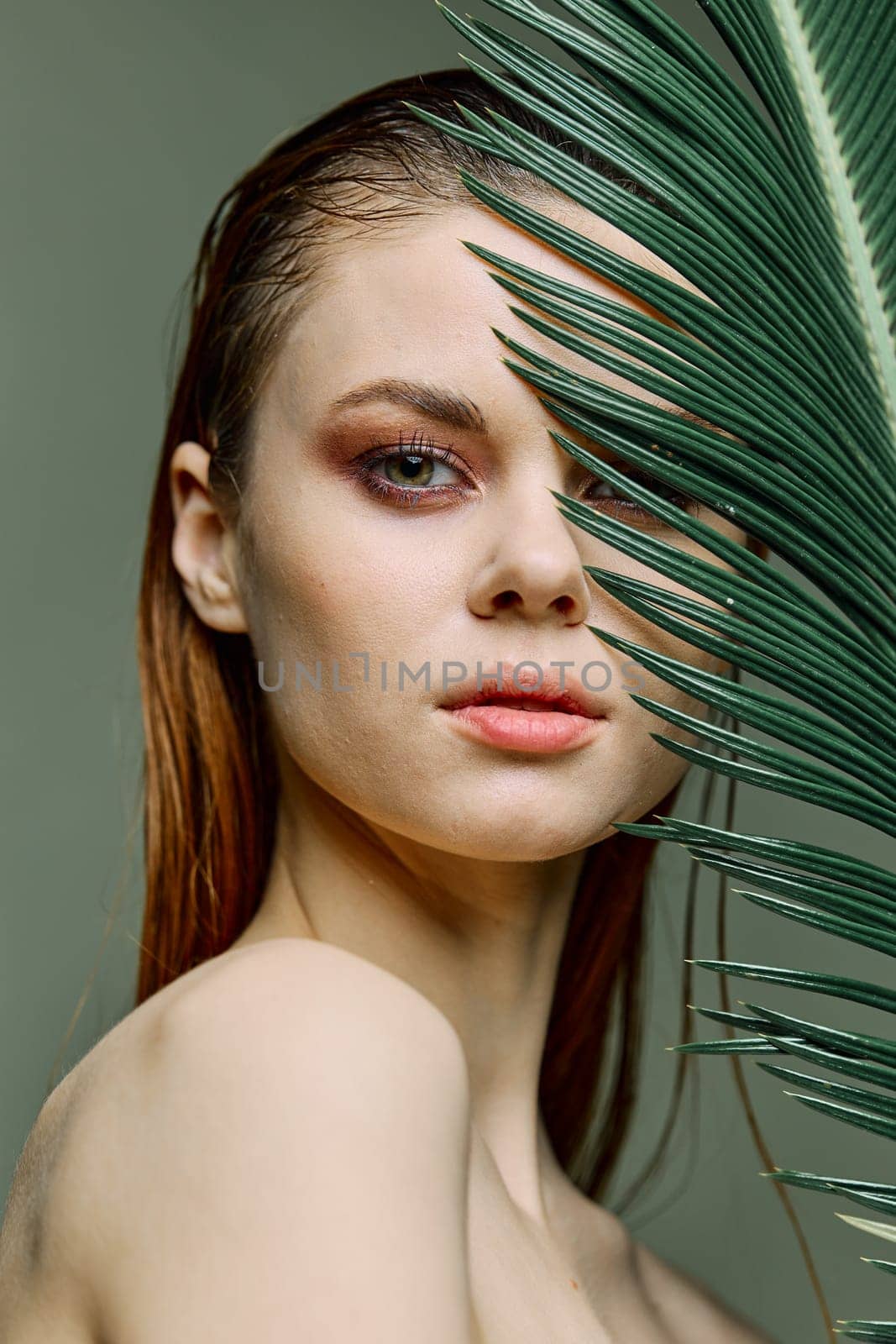 a close beauty portrait of a beautiful woman standing holding a tropical palm leaf in her hand, bringing it to her face, looking into the camera. Vertical photo without retouching of problem skin. High quality photo