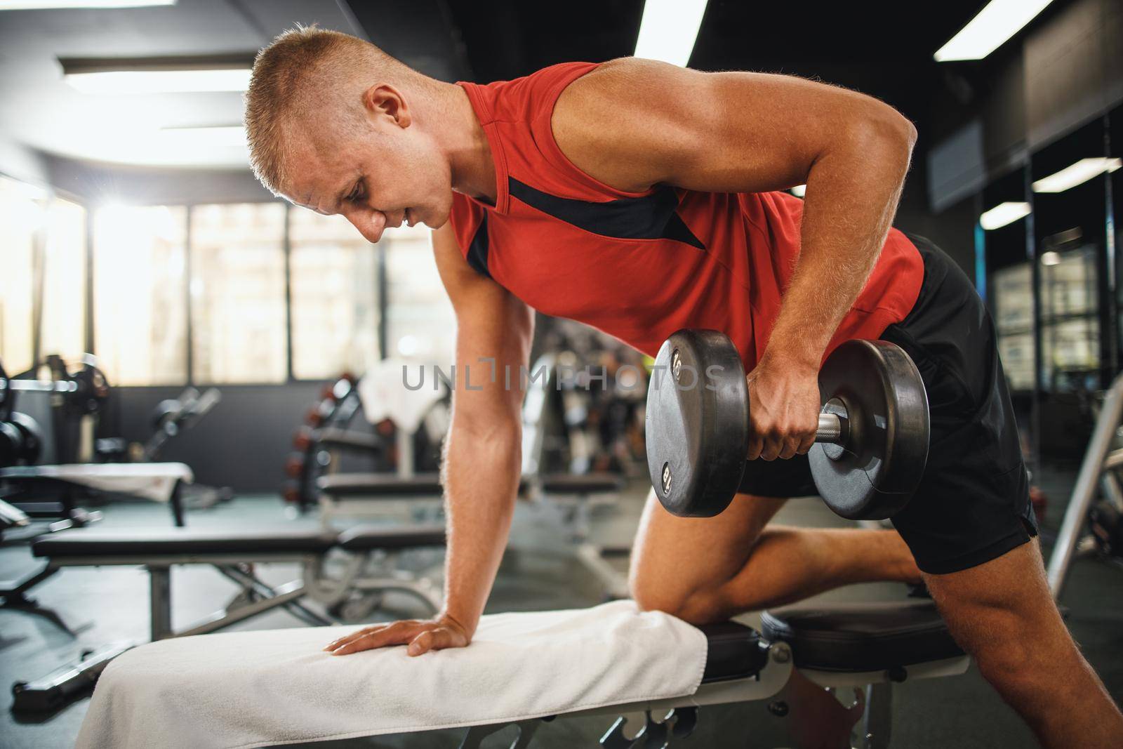 Shot of a muscular guy in sportswear working out at the hard training in the gym. He is pumping his back muscle with dumbbell.