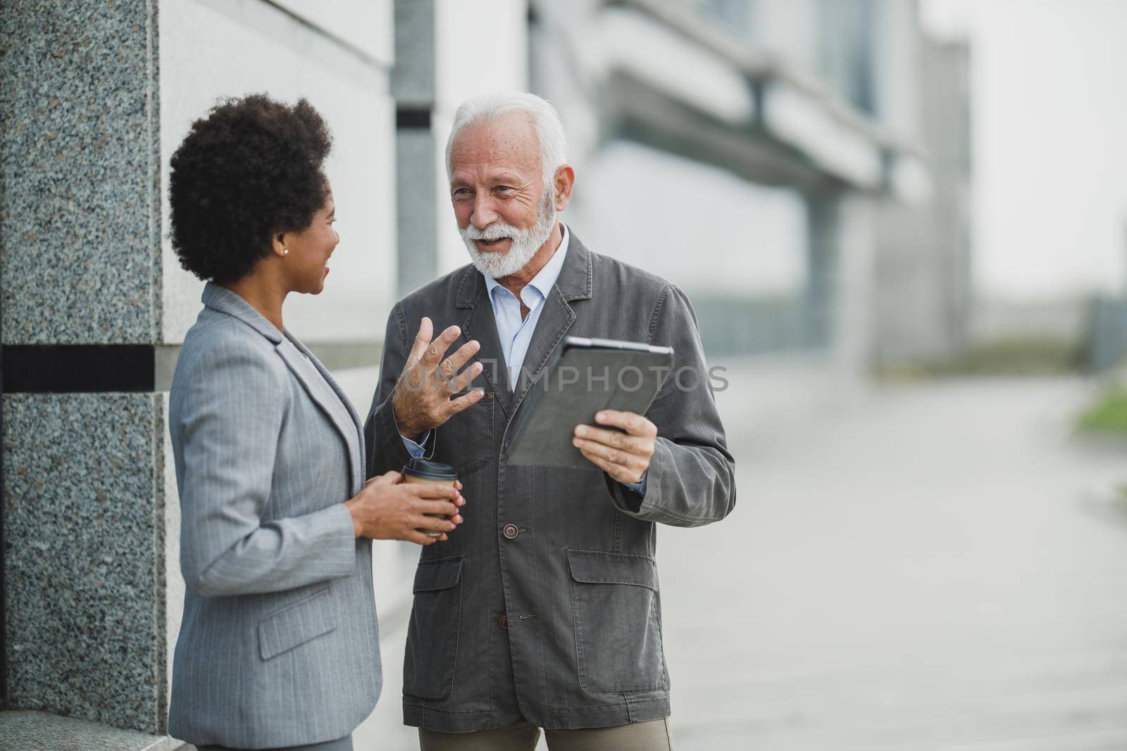 Shot of successful senior businessman and his black female colleague talking and using digital tablet during a coffee break in front of the office building.