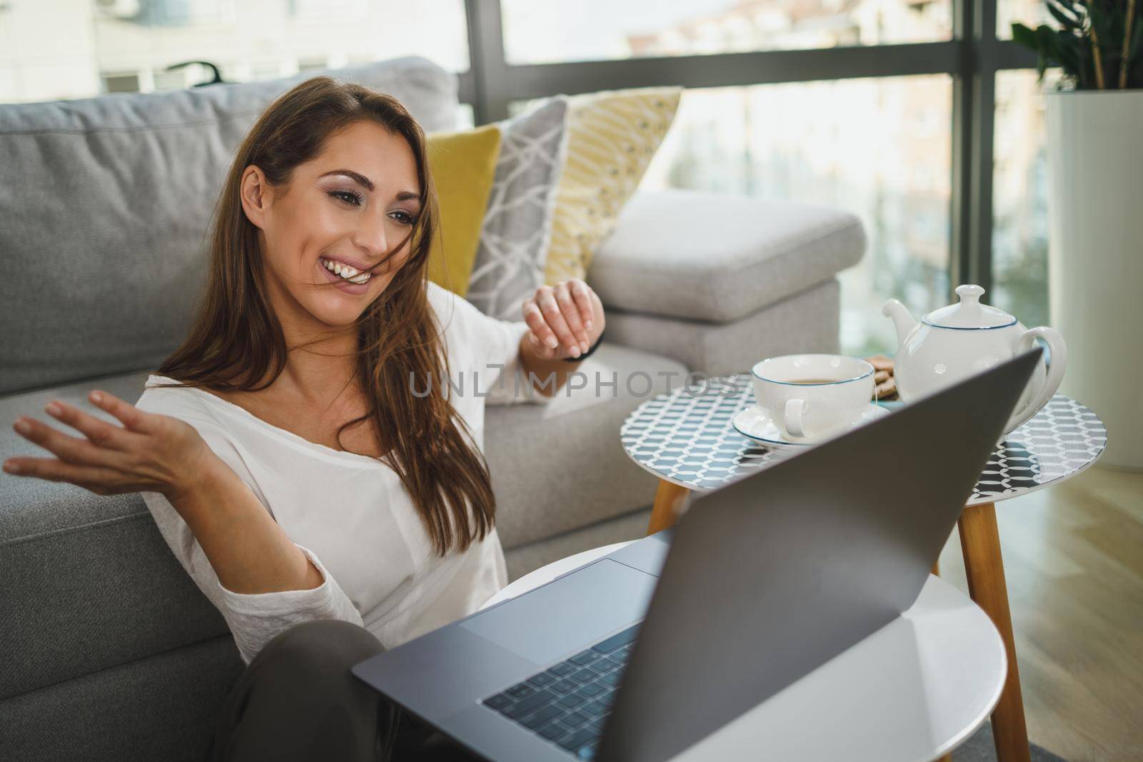 Shot of an beautiful young woman sitting on carpet in her living room and making video call on laptop while having leisure time at her home.