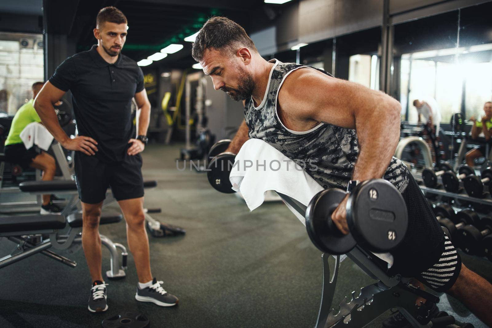 Shot of a muscular guy in sportswear working out with personal trainer at the gym. He is pumping up back muscle with dumbbell.