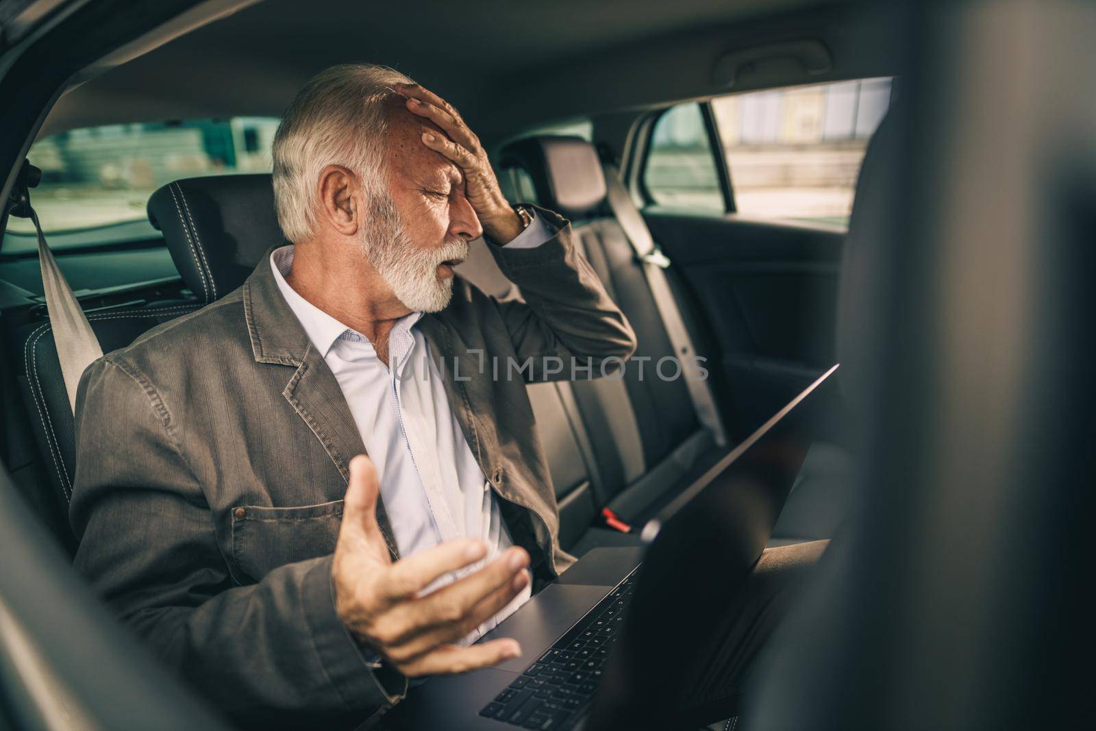 Shot of a stressed senior man using laptop while sitting in the backseat of a car during his business commute.