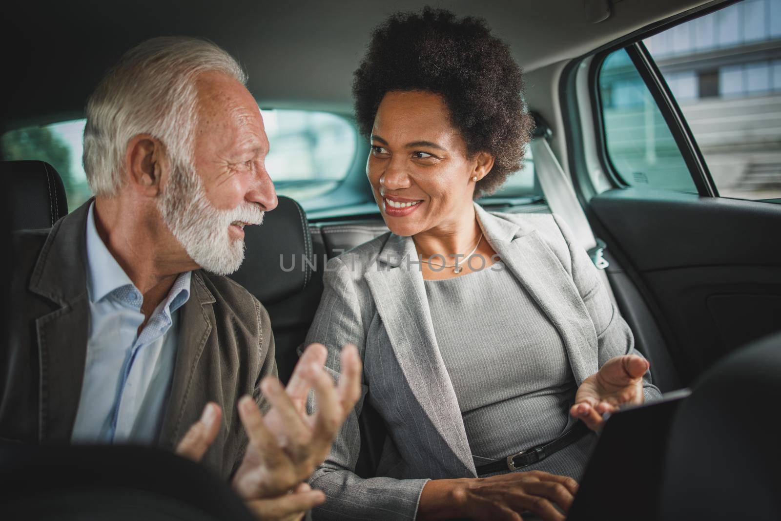 Shot of two successful multi-ethnic people using laptop and having a discussion while sitting in the backseat of a car during their business commute.