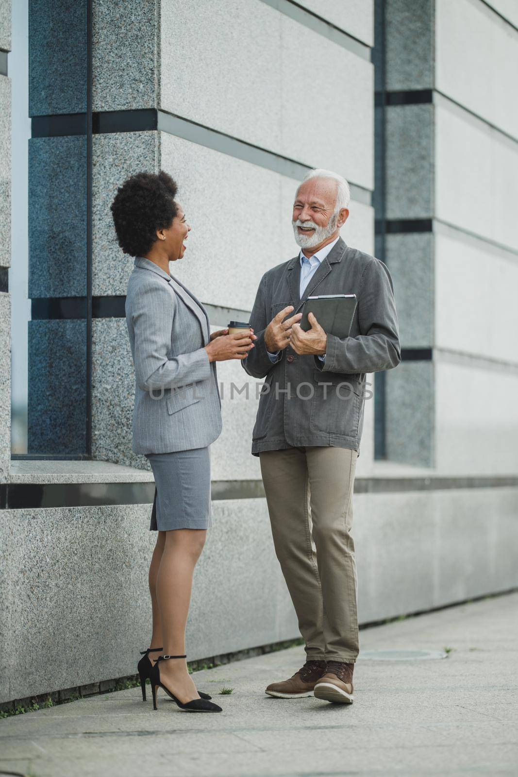 Shot of successful senior businessman and his black female colleague having fun during a coffee break in front of the office building.
