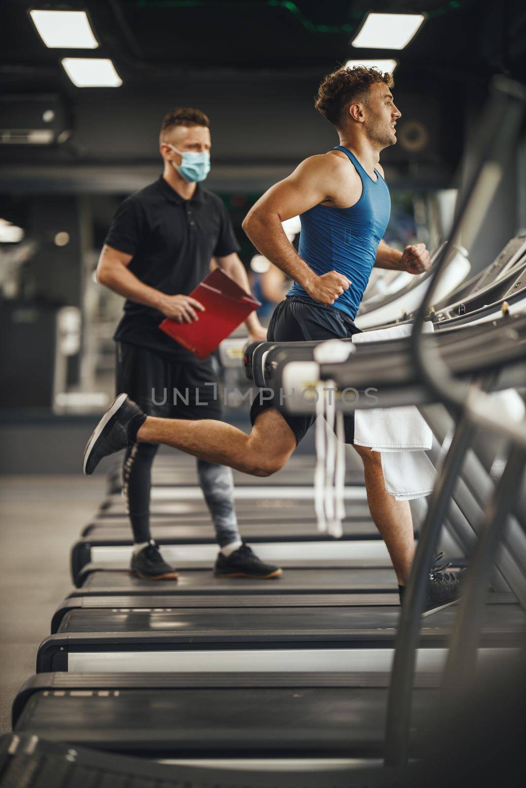 Shot of an muscular young man running on the treadmill at the gym. He is working out with personal trainer.