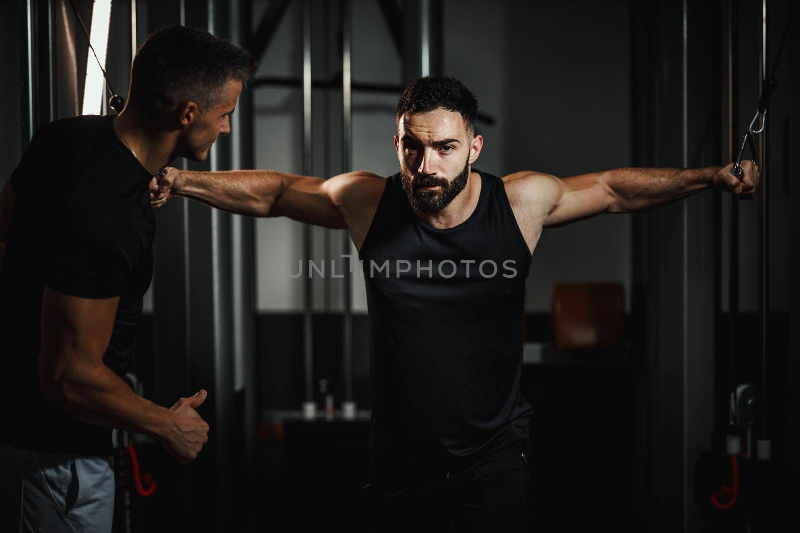 Shot of a muscular guy in sportswear working out with personal trainer at the gym. He is doing hard exercises for chest on cable machine during a strength training.