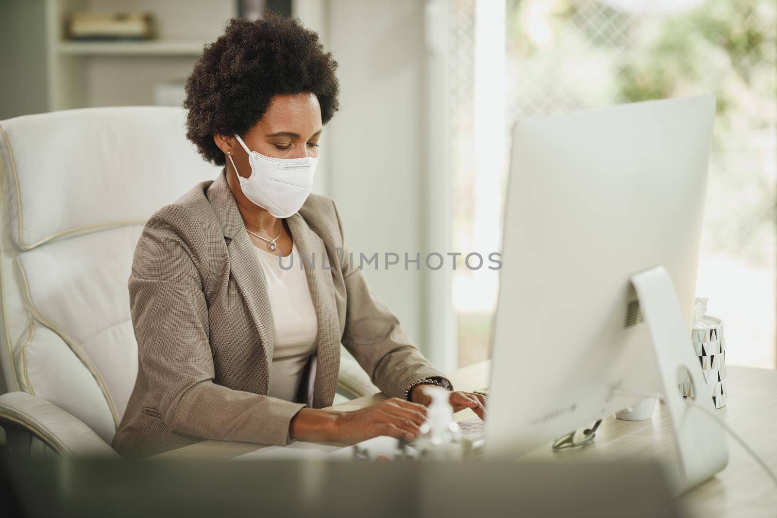 Shot of an African businesswoman with protective N95 mask sitting alone in her office and working on computer during corona virus pandemic.