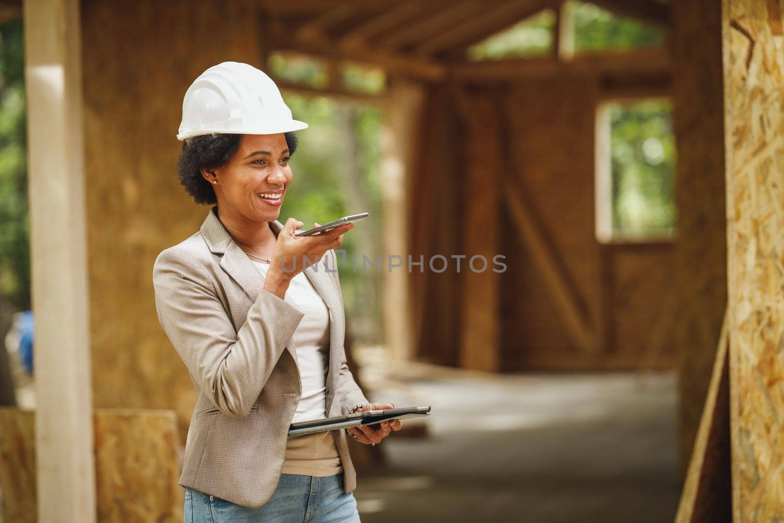 Shot of an African female architect with white helmet using a smartphone and checking construction site of a new wooden house.