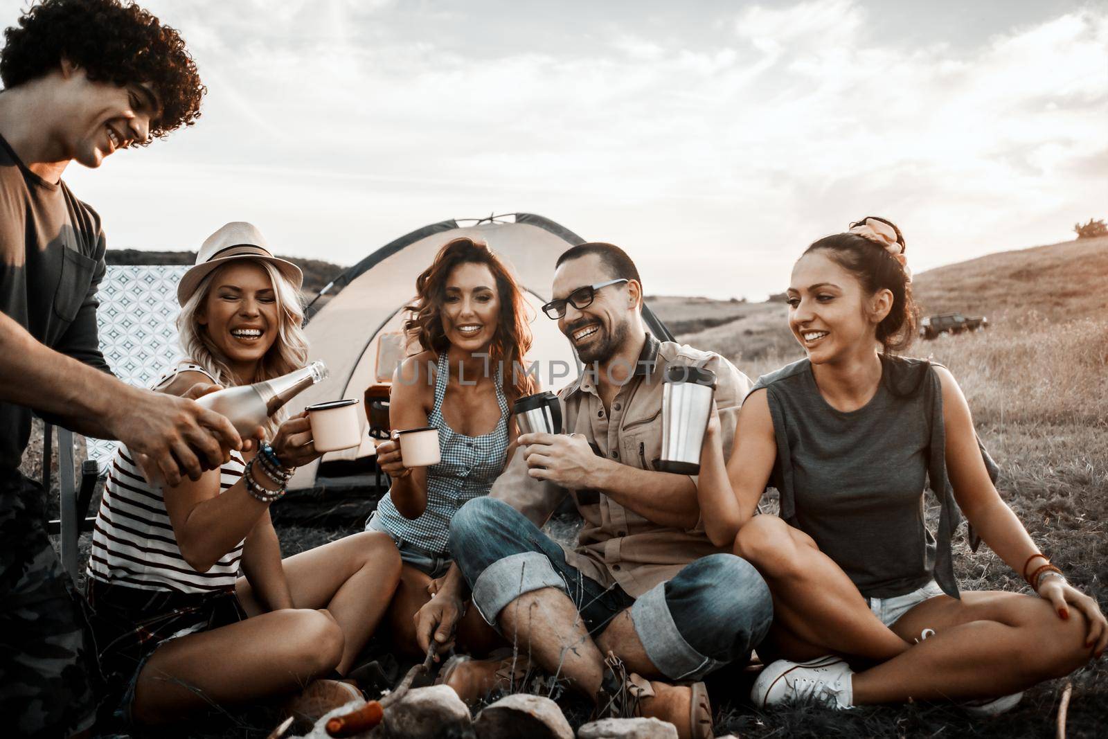 A group of young smiling friends enjoying drink by the fire while on a camping trip.