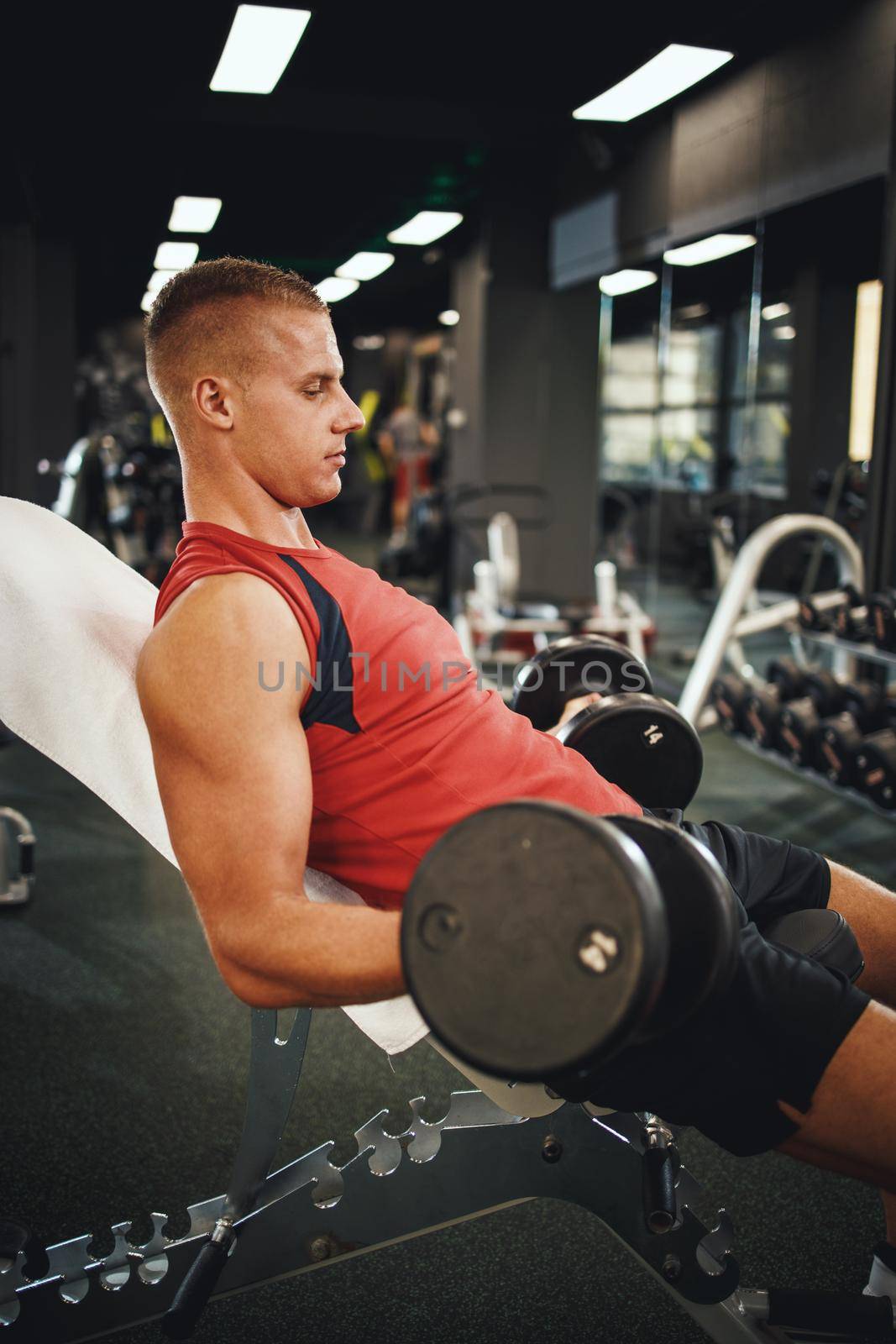 Shot of a muscular guy in sportswear working out at the hard training in the gym. He is pumping his biceps muscle with dumbbell.