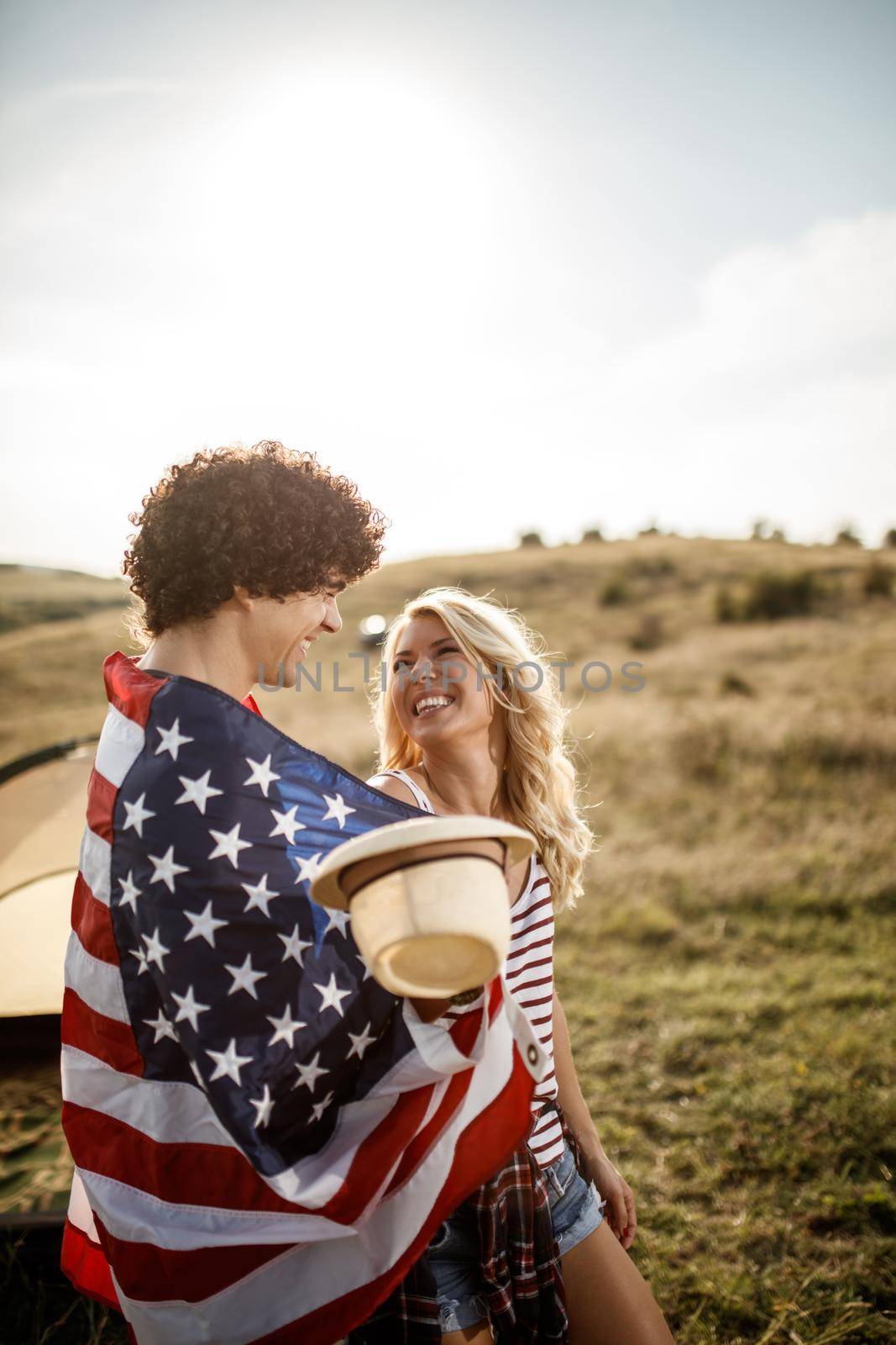 An attractive couple in love wrapped in american flag enjoying in front a tent at the campsite.