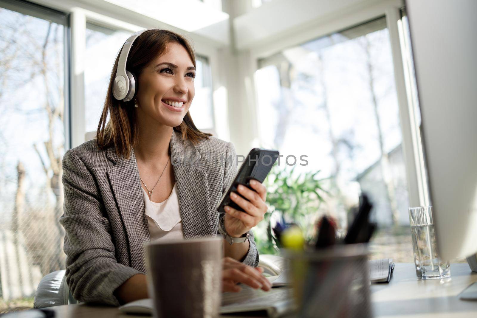 A smiling business woman with headphones using smartphone while working at computer in her home office.
