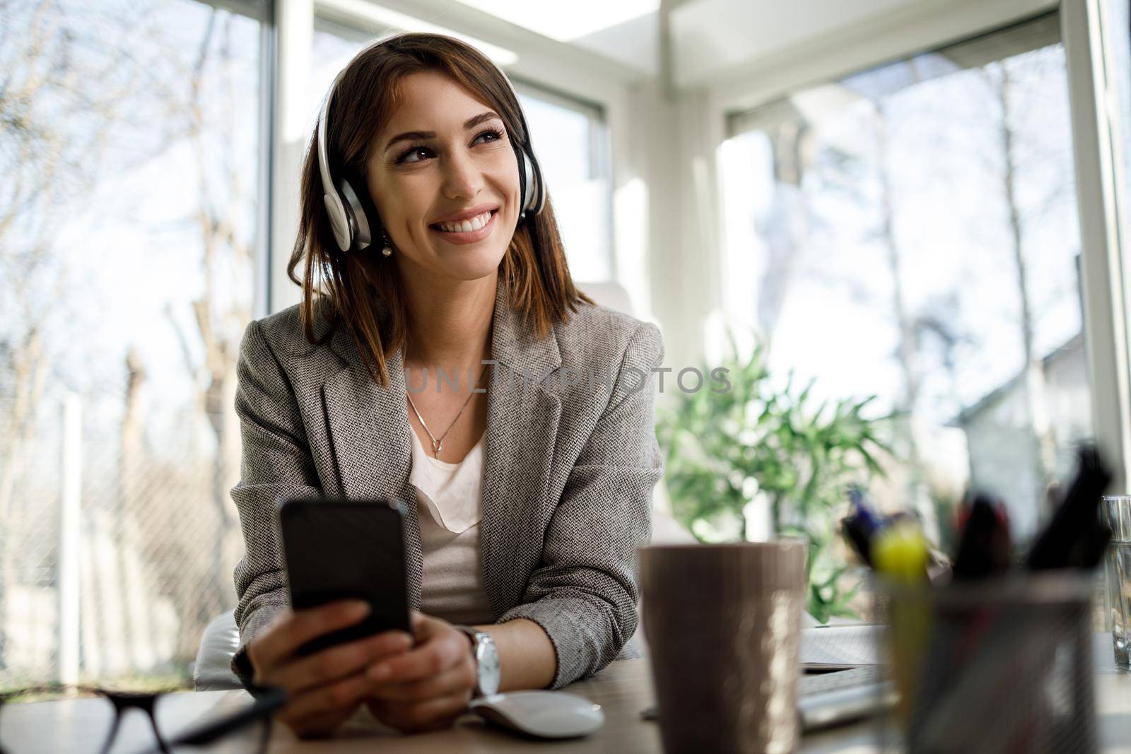A smiling pensive woman using smartphone while working in her home office.