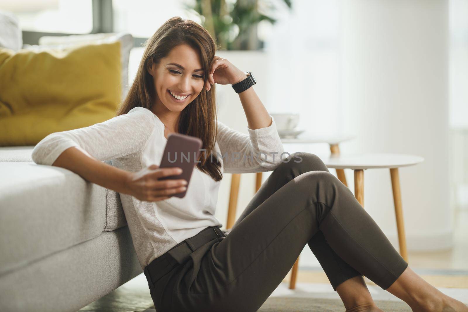Beautiful, young woman sitting next to the window, having leisure time, holding a smart phone and surfing social media at her home.