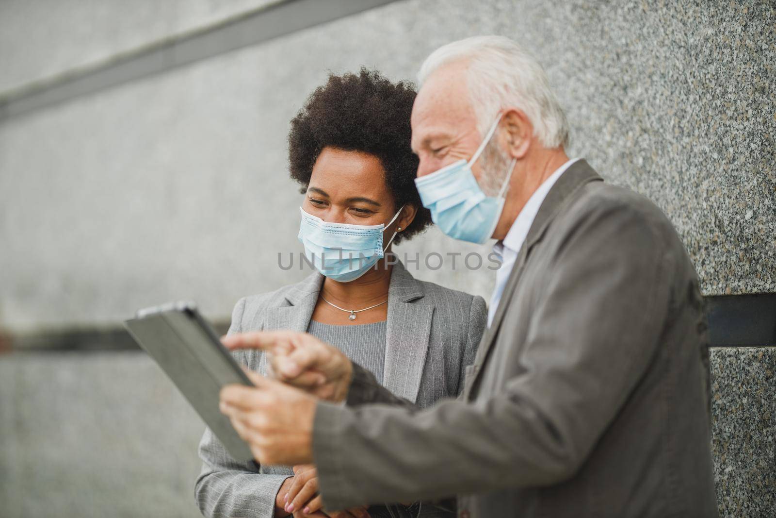 Shot of successful black businesswoman and her senior male colleague with protective mask using digital tablet and having a discussion while standing against a wall of corporate building during COVID-19 pandemic.
