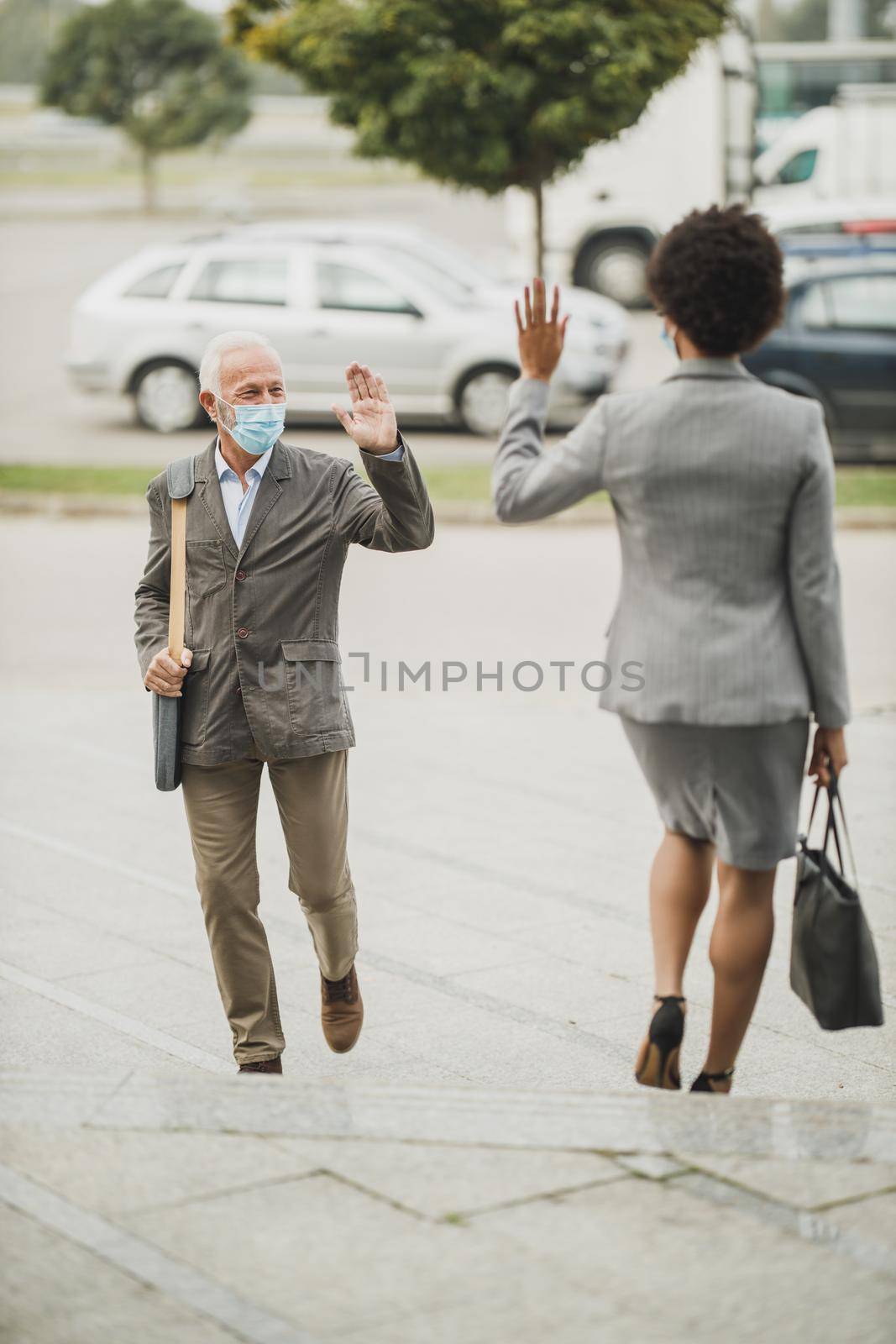 Shot of successful senior businessman and his black female colleague using protective mask and greeting with wave hands during COVID-19 pandemic to avoid handshakes.