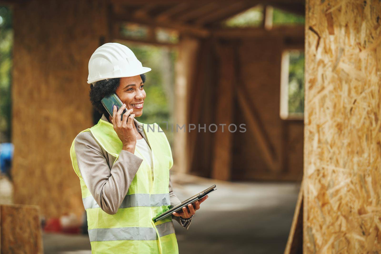 Shot of an African female architect using a smartphone and checking construction site of a new wooden house. She is wearing protective workwear and white helmet.