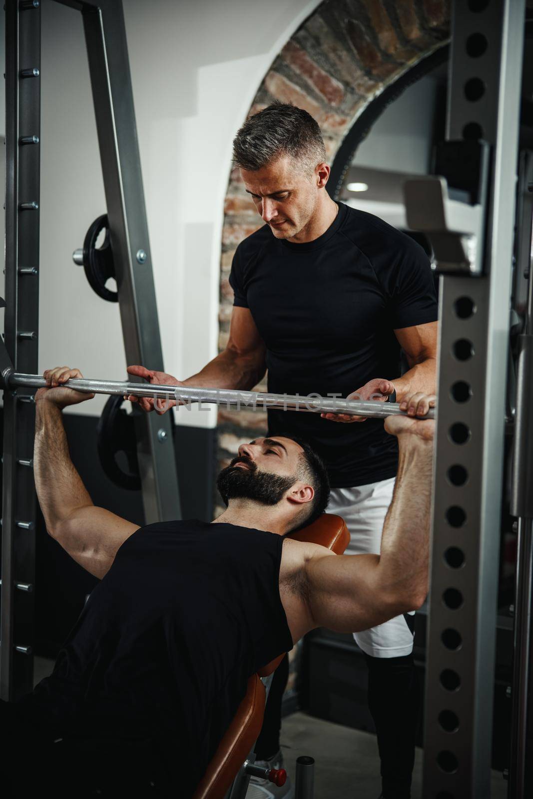 Shot of a muscular guy in sportswear working out with personal trainer at the gym. He is pumping up chest muscule during a hard ttraining.