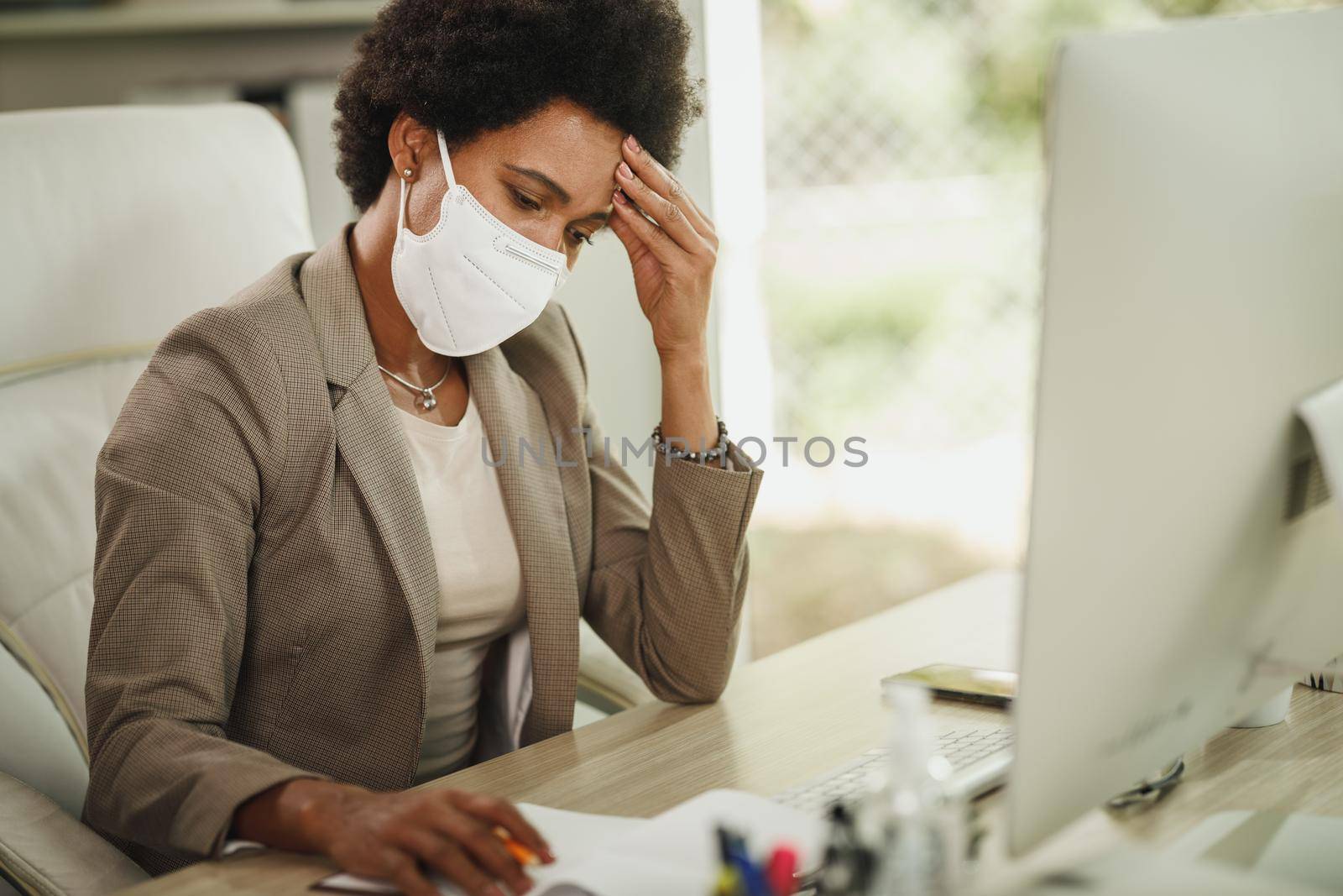 Shot of a worried African businesswoman with protective N95 mask sitting alone in her office and working on computer during COVID-19 pandemic.