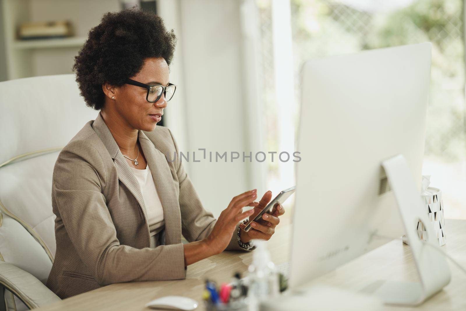 Shot of an African businesswoman using smartphone while working at desk in her home office during corona virus pandemic.