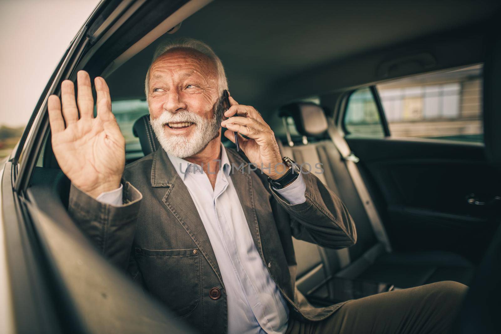 Shot of a successful senior man talking on a smartphone and waving while sitting in the backseat of a car during his business commute.