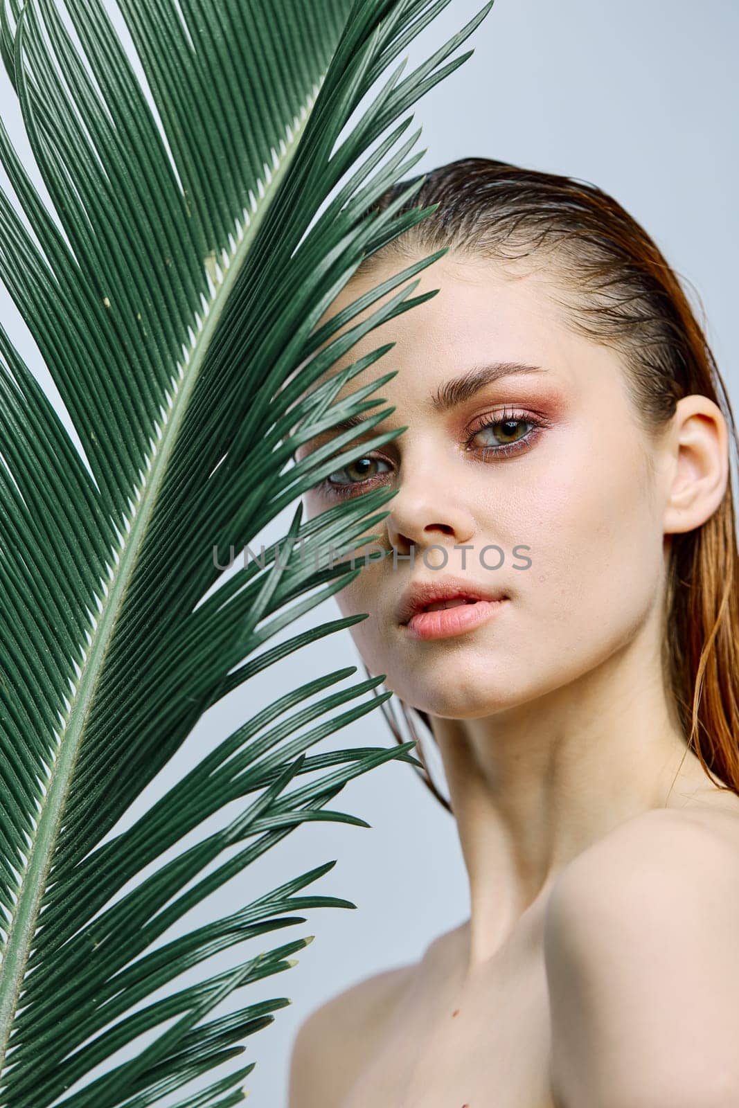 a close beauty portrait of a beautiful woman standing holding a tropical palm leaf in her hand, bringing it to her face. Vertical photo without retouching by Vichizh