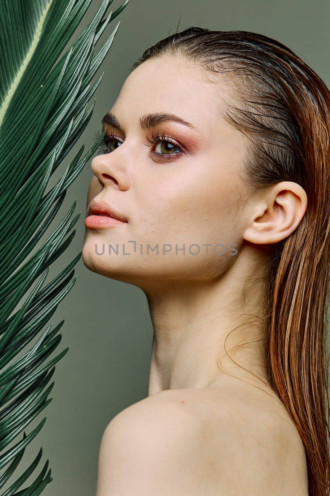 a sophisticated, elegant woman with evening makeup, stands with a green palm leaf, covering part of her face with it. Vertical photo without retouching. High quality photo