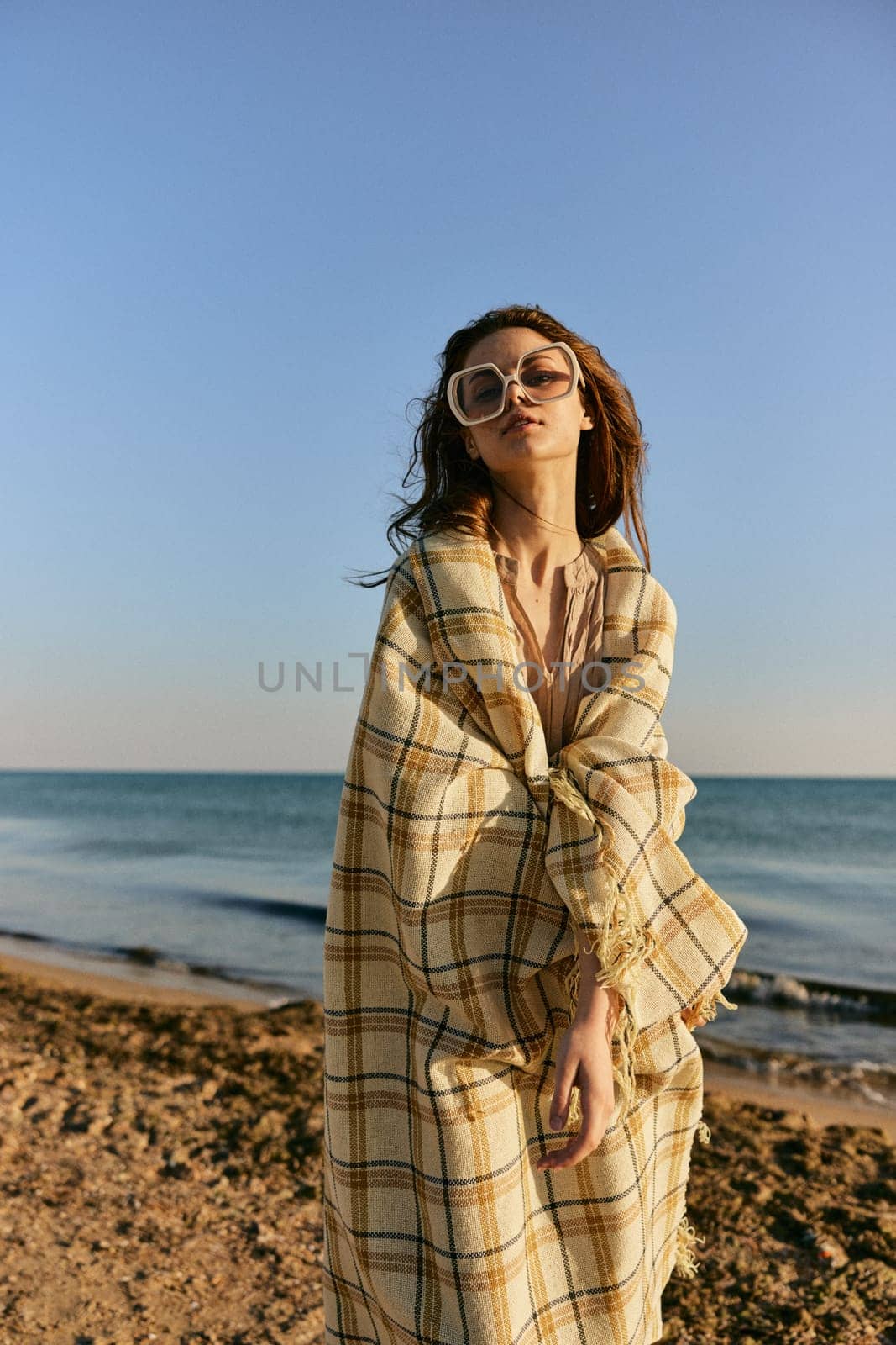 a woman wrapped in a plaid stands on the seashore in bright sunglasses against the background of the sunset sky. High quality photo