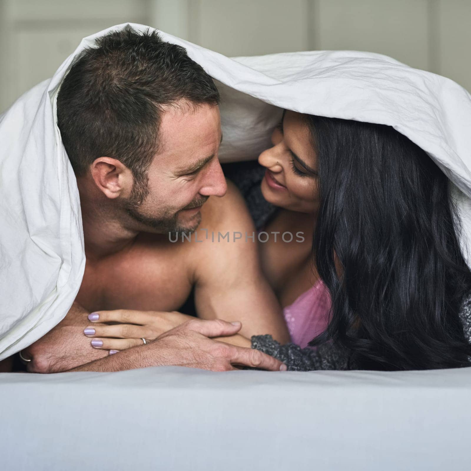 Love under the covers. an affectionate young couple kissing while lying under the covers in bed. by YuriArcurs